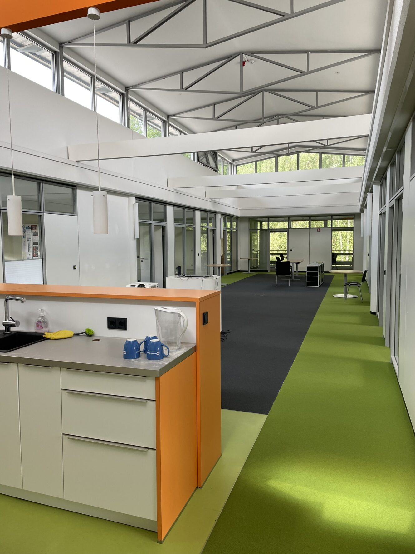 Moderner Büroraum mit grünem Teppich, weißen Wänden, großen Fenstern, einer Küchenzeile im Vordergrund und Schreibtischen und Stühlen im Hintergrund.