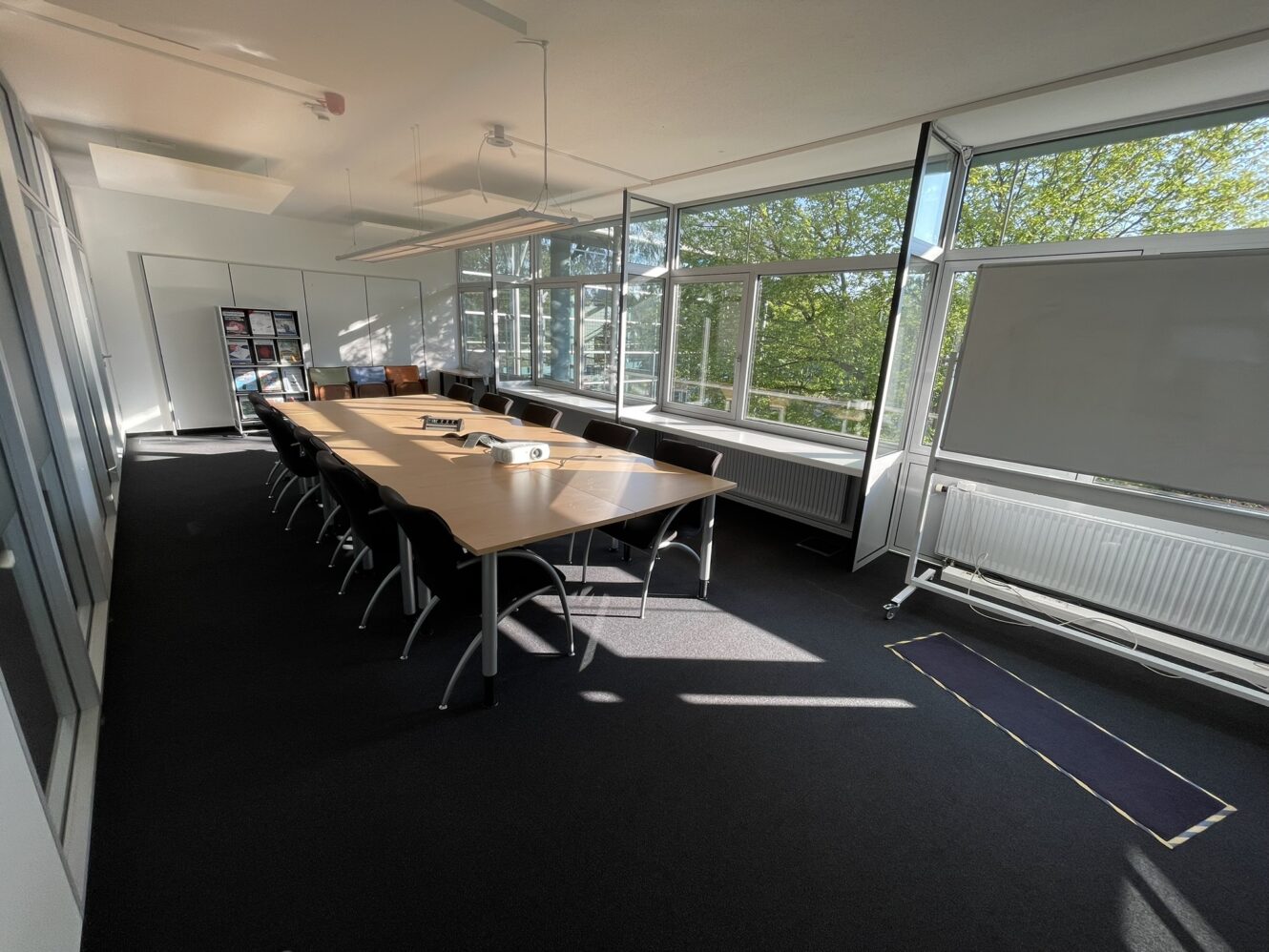 Ein sonnendurchfluteter Konferenzraum mit einem langen Tisch, mehreren Stühlen, einem Whiteboard und großen Fenstern mit Blick auf grüne Bäume.