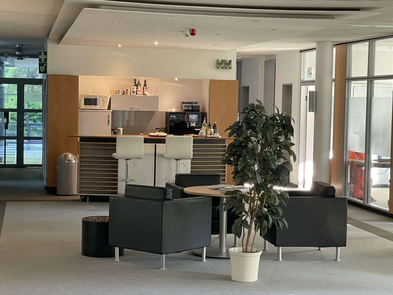 Moderner Büro-Loungebereich mit schwarzen Sesseln, einem runden Tisch, einer Topfpflanze und einem kleinen Küchenbereich mit Barhockern und Geräten im Hintergrund.