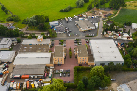 Luftaufnahme eines Industriekomplexes mit Lagerhallen, Bürogebäuden, geparkten Fahrzeugen und umliegenden Grünflächen an einem teilweise bewölkten Tag.