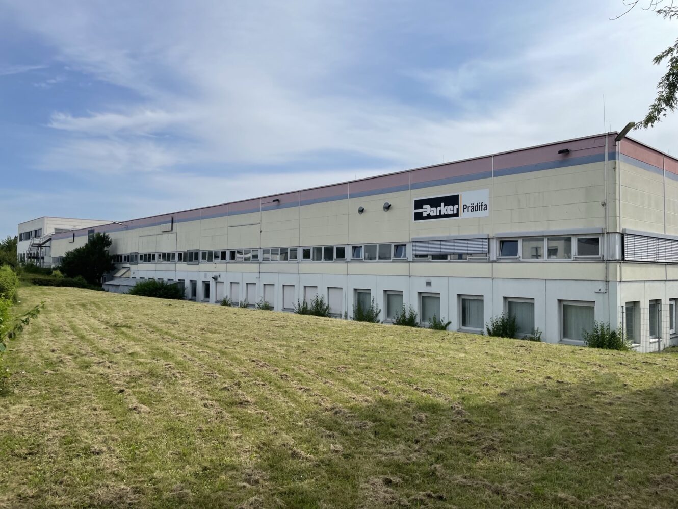Großes Industriegebäude mit Parker Prädifa-Schriftzug, umgeben von einer sauber gemähten Rasenfläche unter einem teilweise bewölkten Himmel.