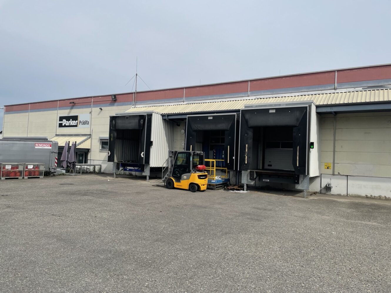 Ein gelber Gabelstapler ist vor den Laderampen eines Lagergebäudes mit Parker-Beschilderung geparkt. Der Bereich scheint bis auf einige Paletten und Container leer zu sein.