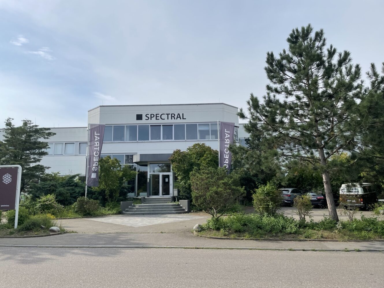 Ein modernes weißes Bürogebäude mit SPECTRAL-Beschilderung, zwei vertikalen Bannern und einem begrünten Eingang, der von Bäumen und Grünflächen umgeben ist.