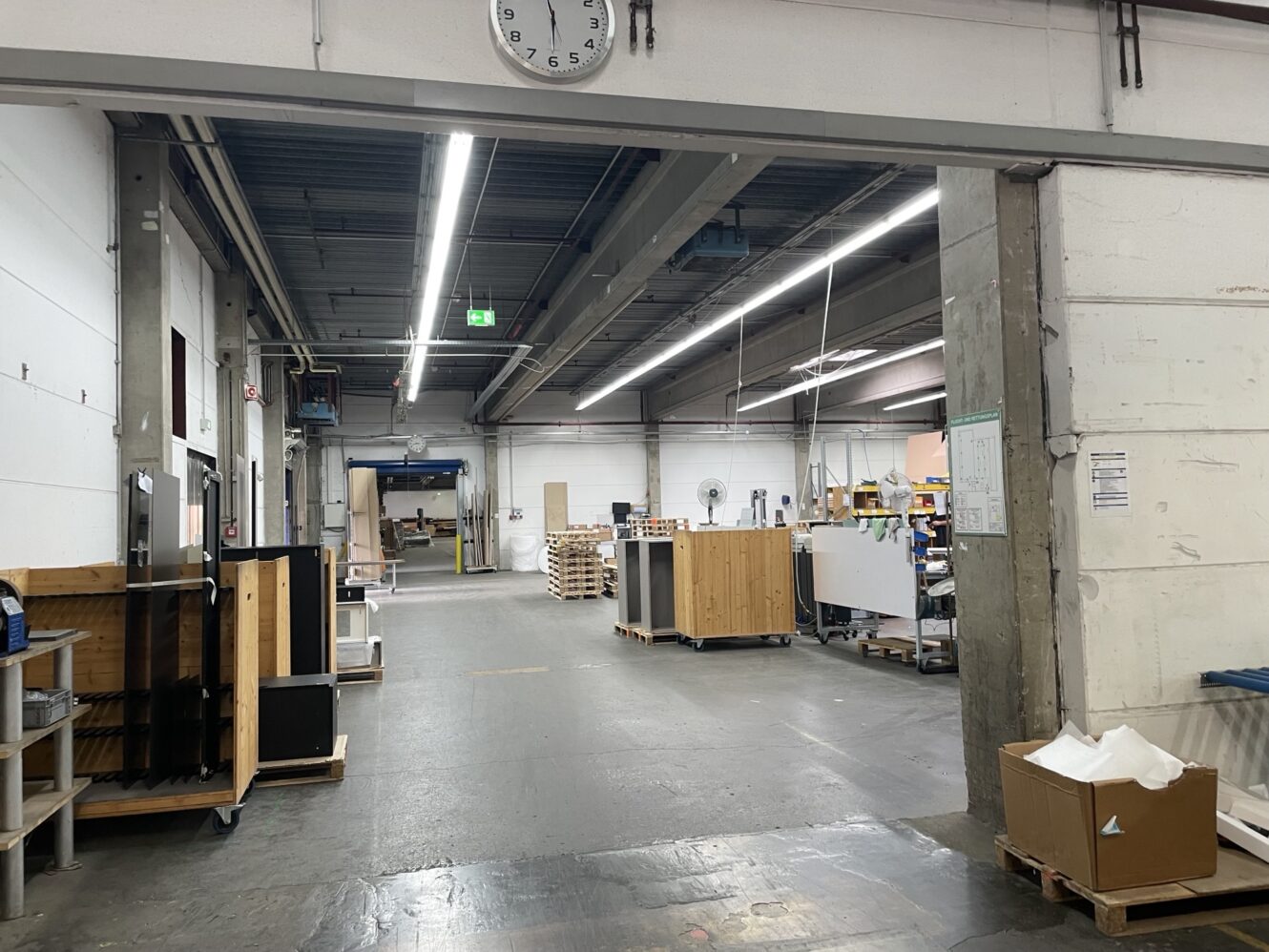Ein geräumiges Lager mit Industrieregalen, Paletten und Geräten unter Leuchtstoffröhren; über dem Eingang ist eine Uhr angebracht.