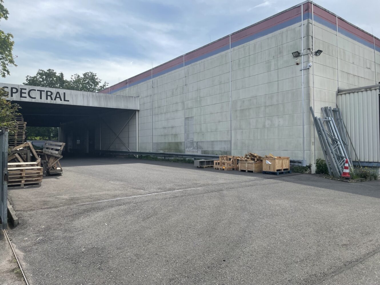 Ein großes Industriegebäude mit SPECTRAL, das teilweise auf einem Schild zu sehen ist, Holzpaletten, die draußen gestapelt sind, und Überwachungskameras, die an der Ecke angebracht sind.
