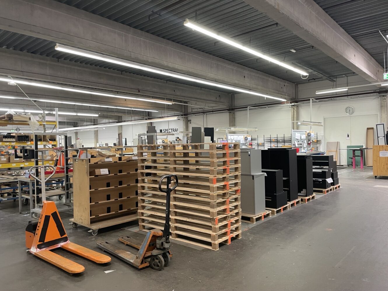 Industrielager mit Holzpaletten, Metallwagen und verschiedenen Geräten und Materialien, die in einem geräumigen, gut beleuchteten Bereich übersichtlich angeordnet sind.