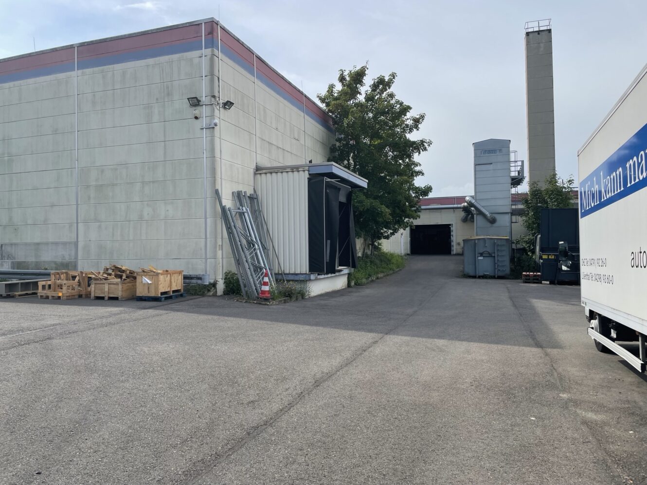 Außenansicht einer Industrieanlage mit Betonwänden, Transportkisten, Metallrohren, Überwachungskameras, einem Baum und einem rechts geparkten weißen Lastwagen.