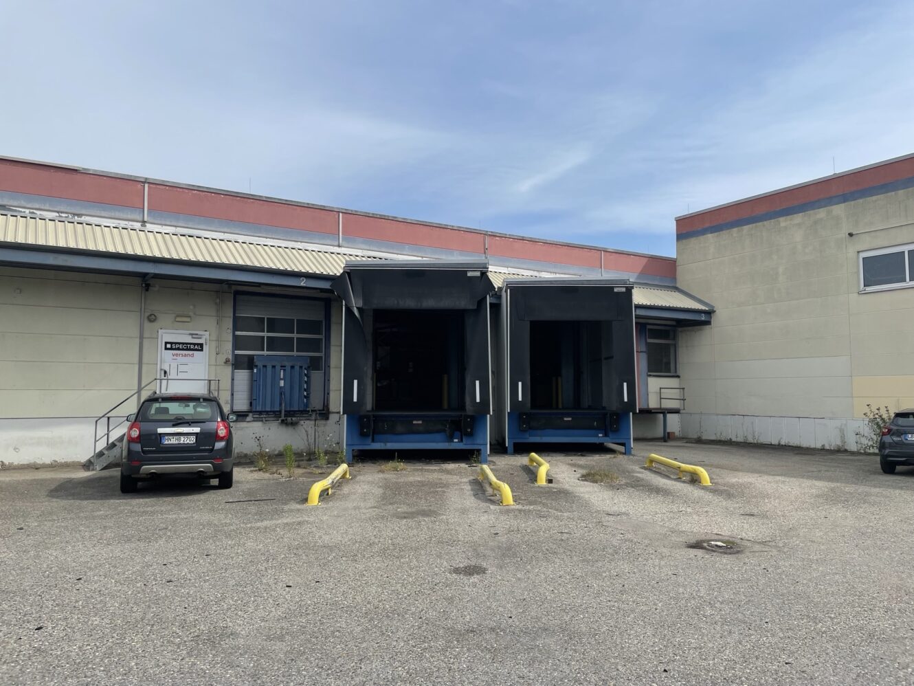 Zwei Laderampen mit erhöhten Plattformen an der Rückseite eines Industriegebäudes; in der Nähe parkt ein schwarzes Auto auf einem gepflasterten Parkplatz mit gelben Schranken.