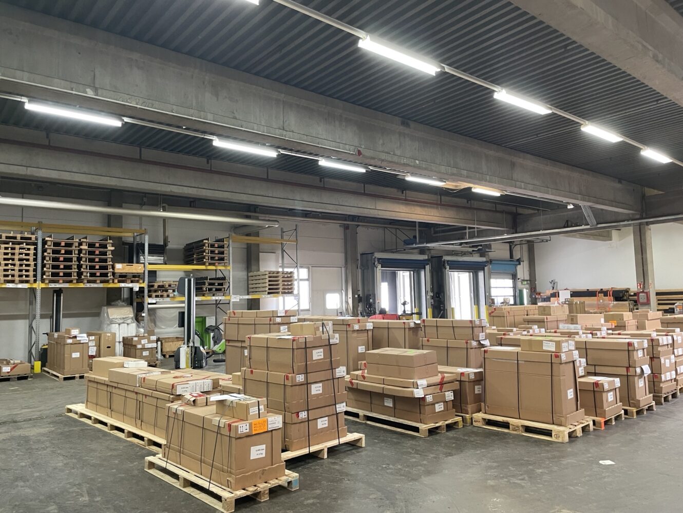 Großes Lagerhaus mit Stapeln von Kartons auf Holzpaletten, Regalen an der Rückwand und Laderampen im Hintergrund.