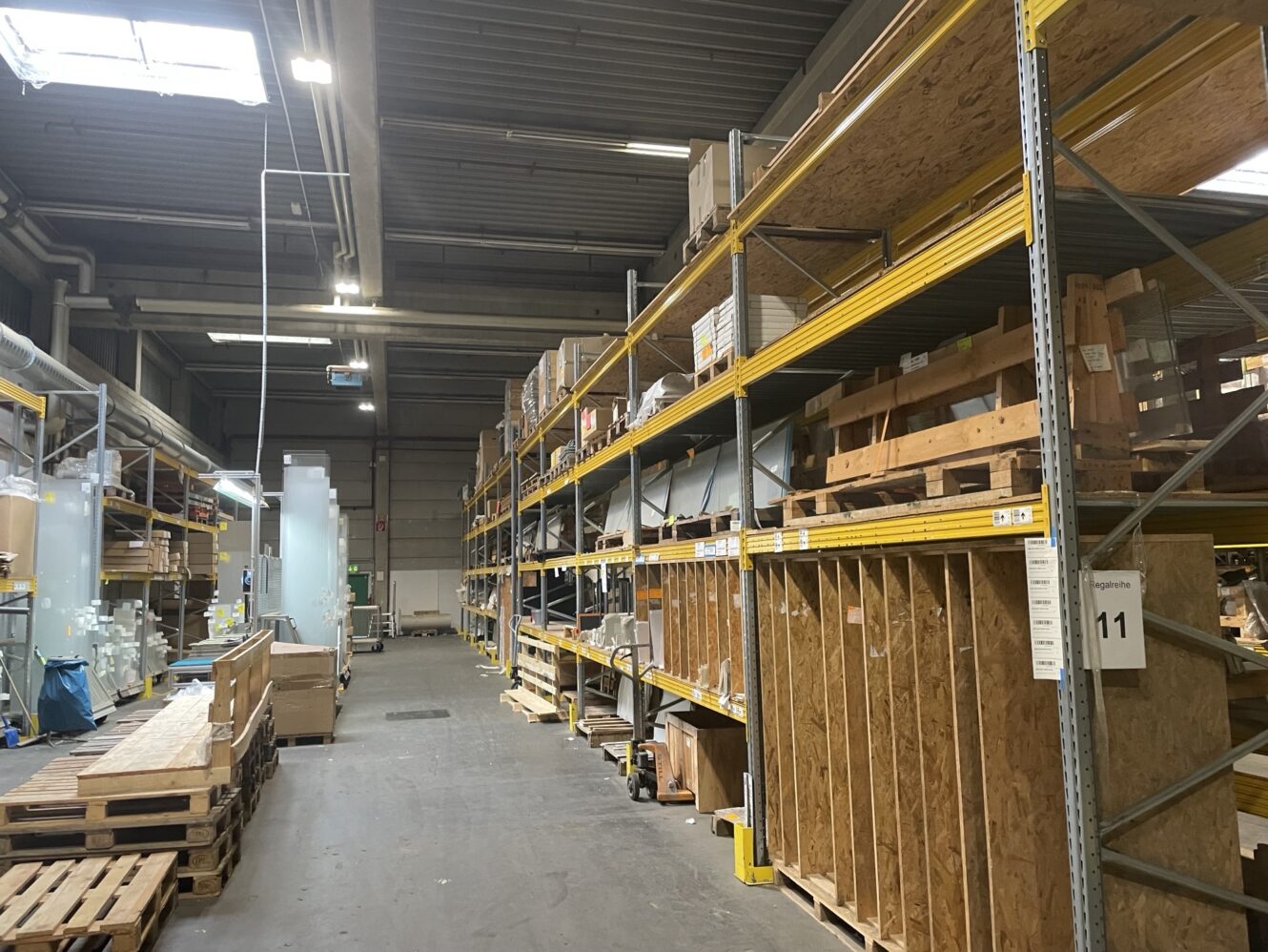 Industrielager mit hohen Metallregalen mit Holz- und Metallmaterialien, leeren Paletten auf dem Boden und heller Deckenbeleuchtung.