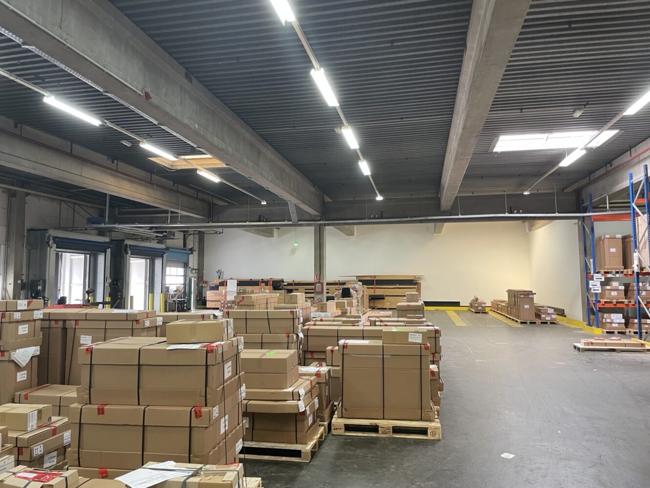 Ein Lagerhaus mit Paletten gestapelter Pappkartons, einige davon beschriftet und in Plastik eingewickelt, unter hellen Oberlichtern.
