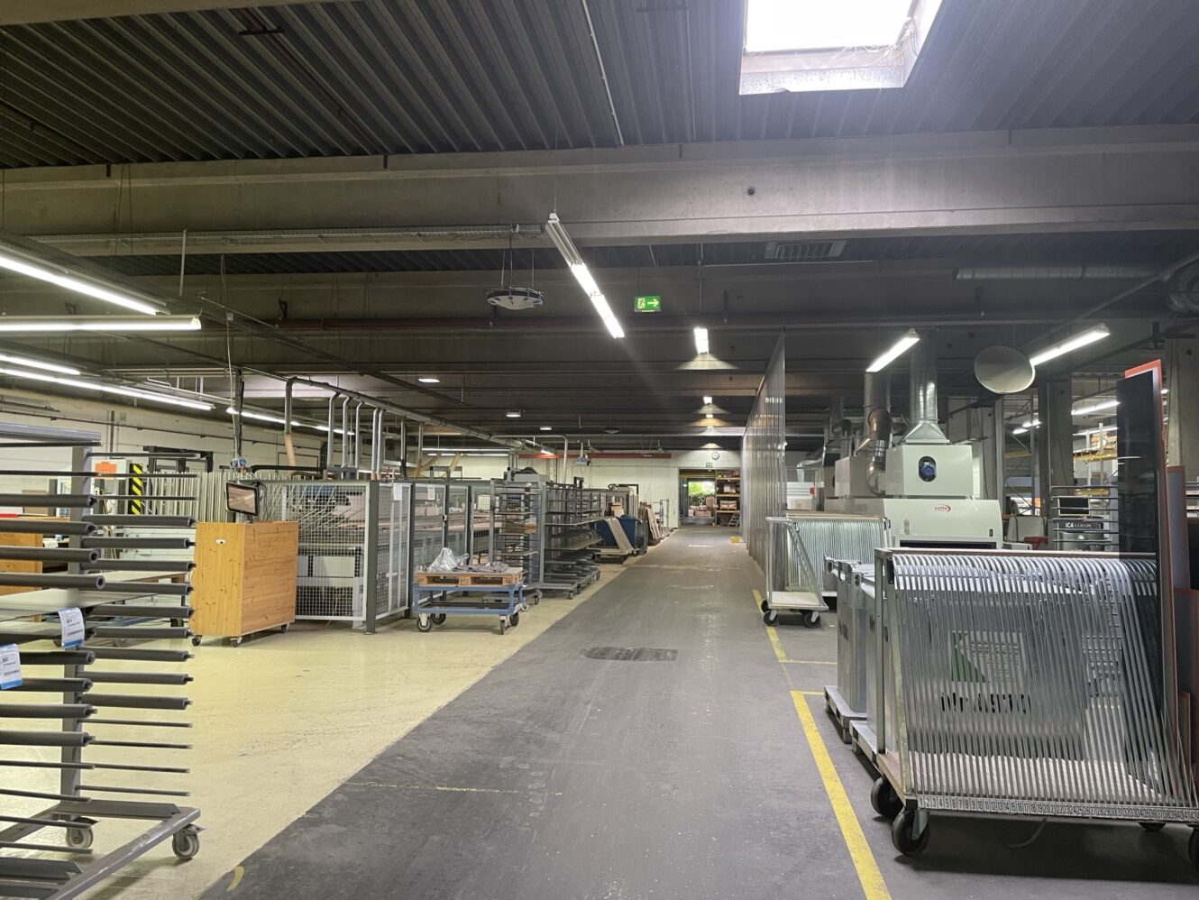 Ein geräumiges Industrielager mit Metallregalen, Wagen und Maschinen unter Leuchtstoffröhrenbeleuchtung. An der Decke sind ein Oberlicht und Lüftungskanäle zu sehen.