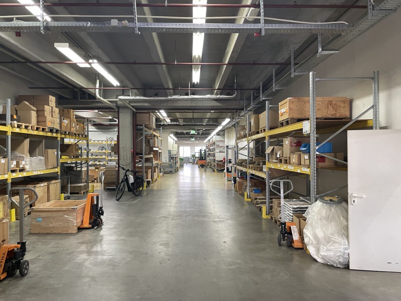 Breiter Gang in einem Lagerhaus mit Regalen auf beiden Seiten, in denen sich Kisten und verschiedene Artikel befinden; außerdem sind Palettenheber und ein Fahrrad zu sehen.