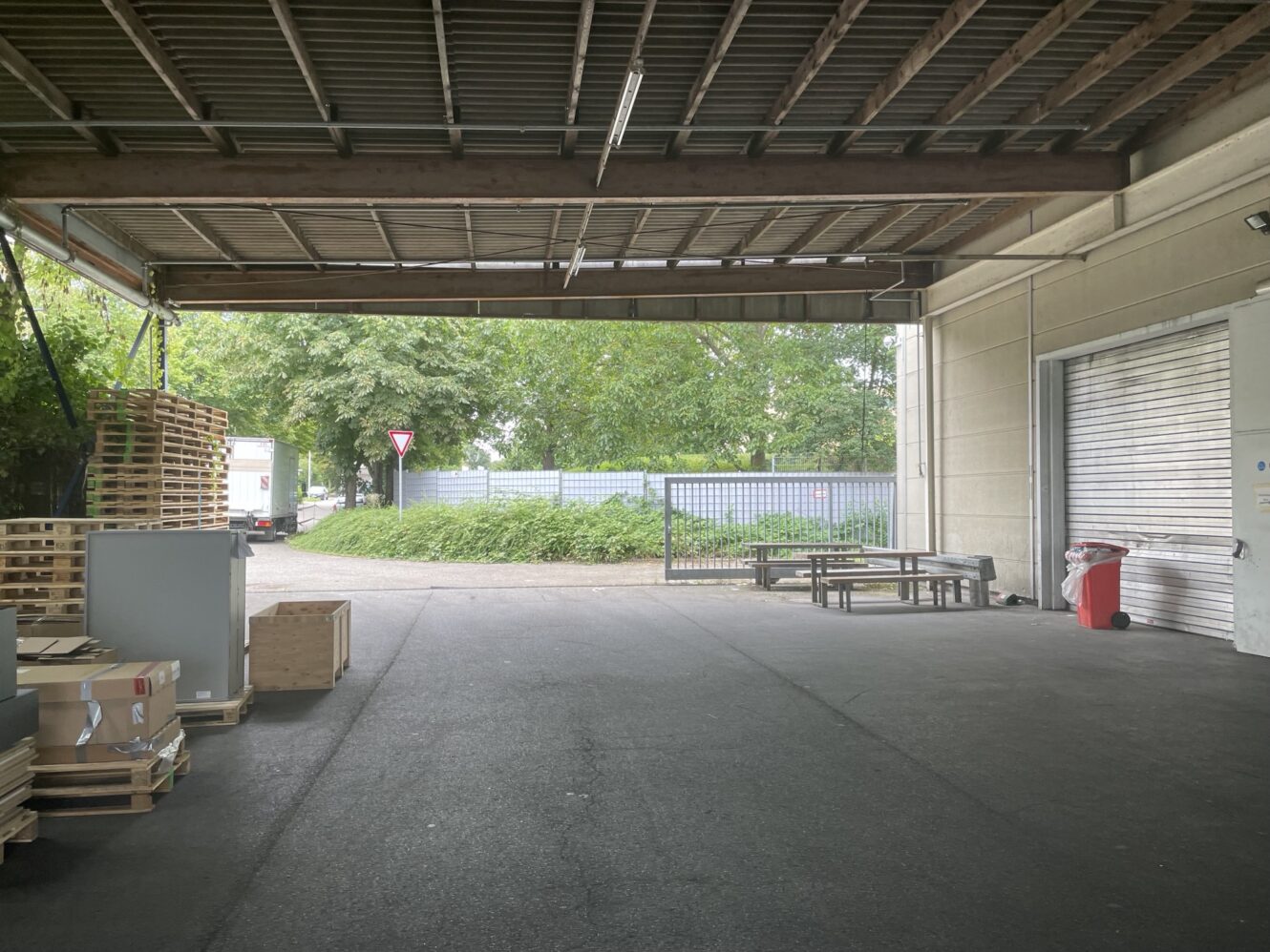 Überdachte Ladefläche mit Paletten, Kisten, Metalltor, Sitzbank, roter Mülltonne und Bäumen im Außenbereich.