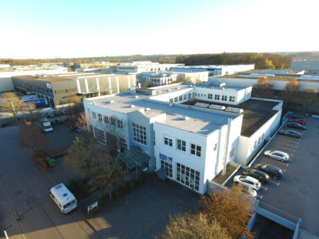 Luftaufnahme eines weißen industriellen Bürogebäudes mit angrenzendem Parkplatz und mehreren geparkten Autos, umgeben von anderen Geschäftsgebäuden und Bäumen.