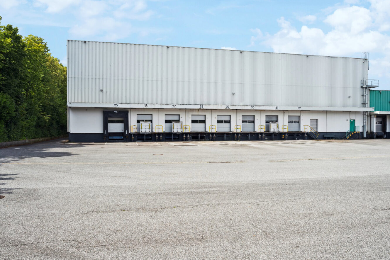 Ein großes weißes Lagergebäude mit mehreren Ladetoren und einem leeren gepflasterten Parkplatz davor.