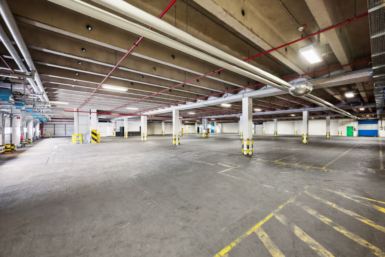 Das Innere eines großen, leeren Industrieparkhauses mit Betonböden, Säulen, oberirdischen Rohren und Leuchtstoffröhren. Gelbe Sicherheitsmarkierungen und Absperrungen sind überall sichtbar.