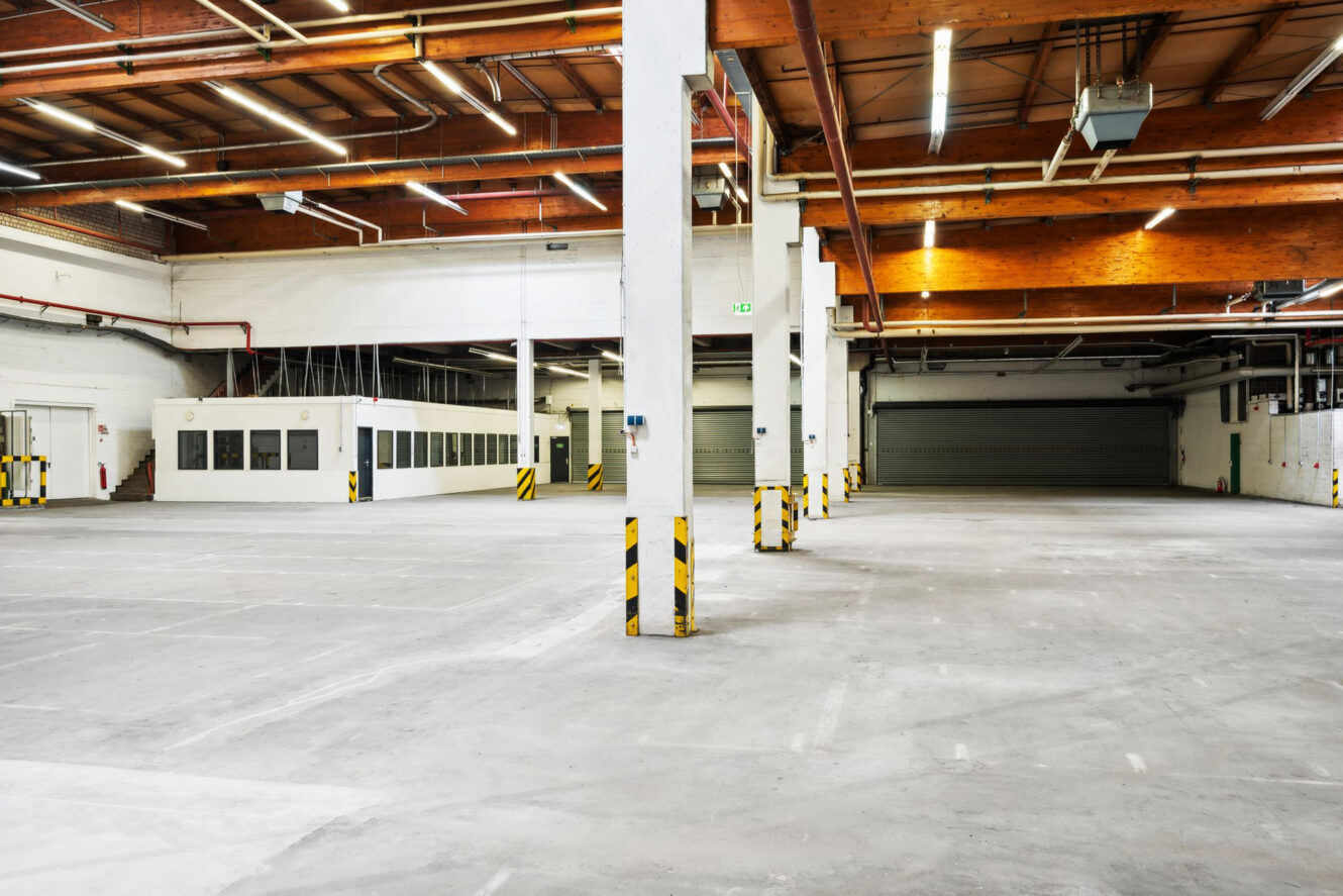 Große, leere Industrielagerhalle mit weißen Wänden, freiliegenden Holzbalken, Säulen mit gelb-schwarzen Markierungen und einer Reihe von geschlossenen Garagentoren.