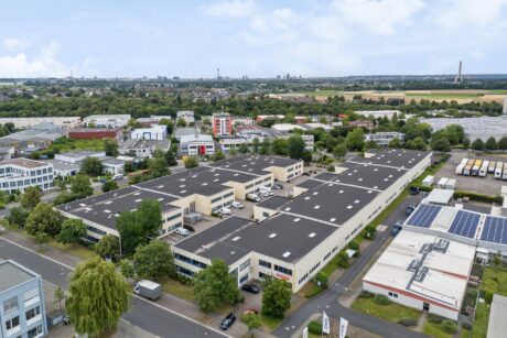 Luftaufnahme eines Industrieparks mit mehreren großen Lagergebäuden, Parkplätzen und umliegenden Grünflächen bei teilweise bewölktem Himmel.