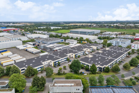 Luftaufnahme eines Industriegebiets mit mehreren Lagergebäuden, geparkten Lastwagen und umliegenden Grünflächen unter einem teilweise bewölkten Himmel.