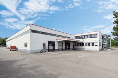 Ein großes, modernes Industrielager mit weißen Außenwänden, roten Verkleidungen, Laderampen und einem angrenzenden Bürotrakt unter blauem Himmel.