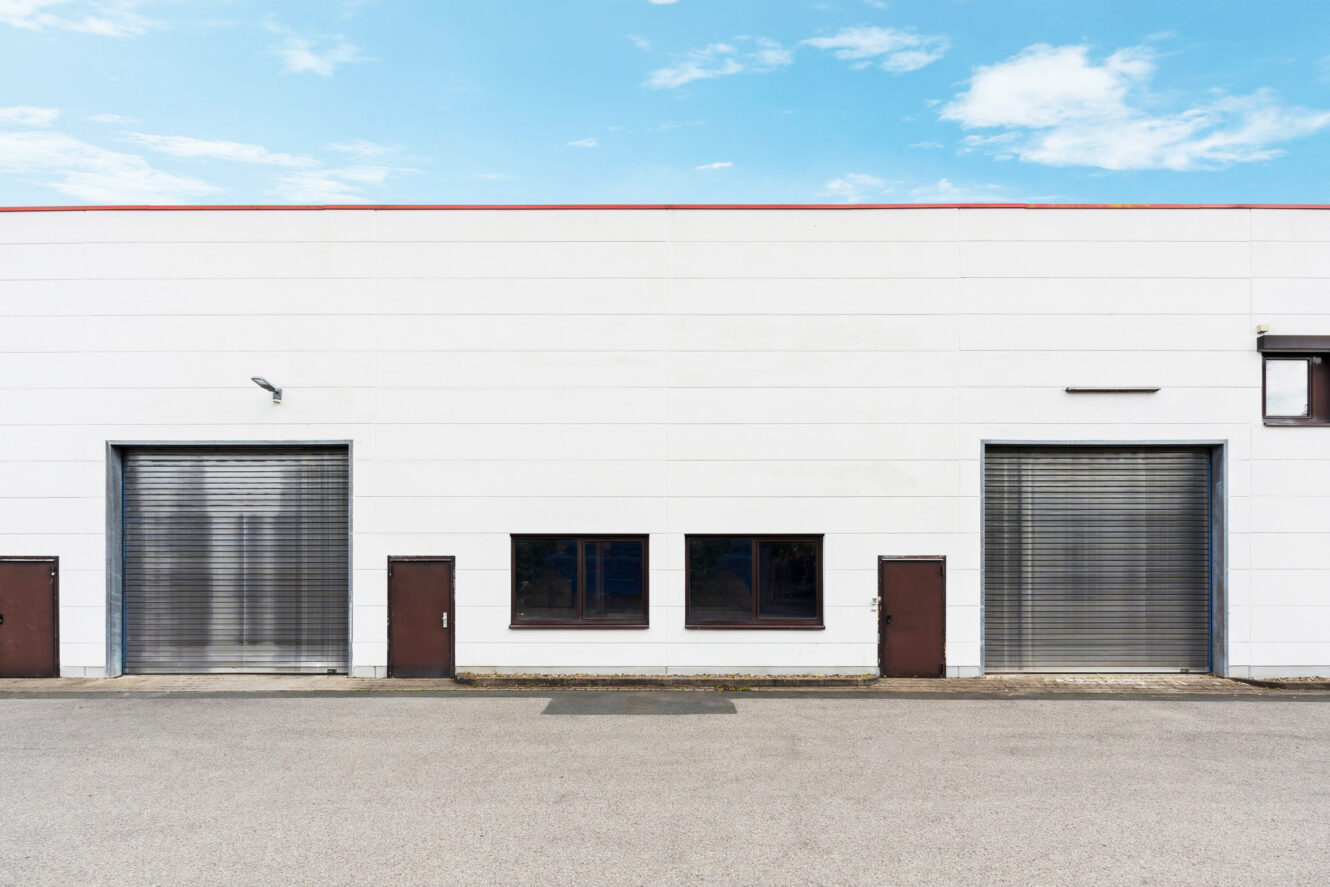 Ein großes Industriegebäude mit schlichter weißer Fassade, zwei großen Metallrolltoren, drei kleineren Türen und mehreren Fenstern vor einem blauen Himmel mit Wolken.