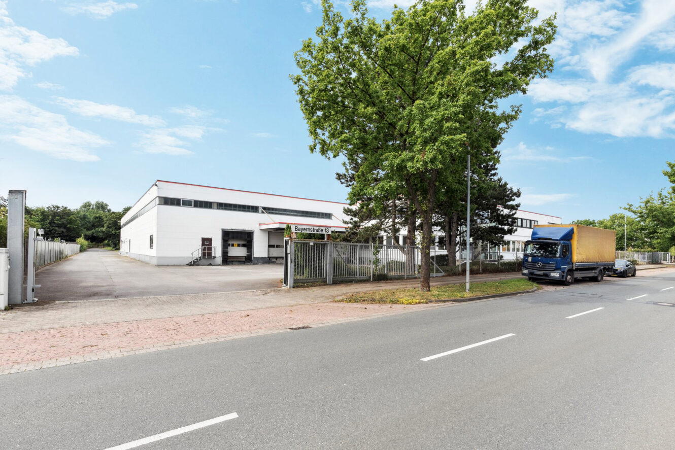 Ein großes weißes Industrielager mit einer Laderampe, einem Metallzaun und einem geparkten blauen Lkw an einer ruhigen Straße unter klarem Himmel.