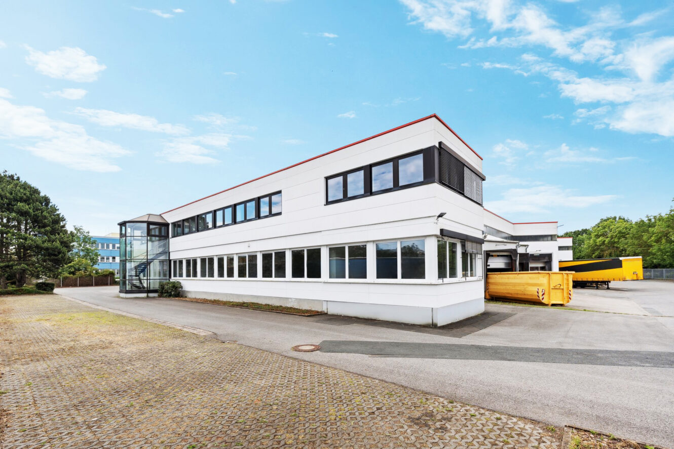 Ein modernes, weißes, zweistöckiges Industriegebäude mit großen Fenstern, einem gläsernen Treppenhaus und gelben Müllcontainern vor dem Hintergrund eines teilweise bewölkten Himmels.