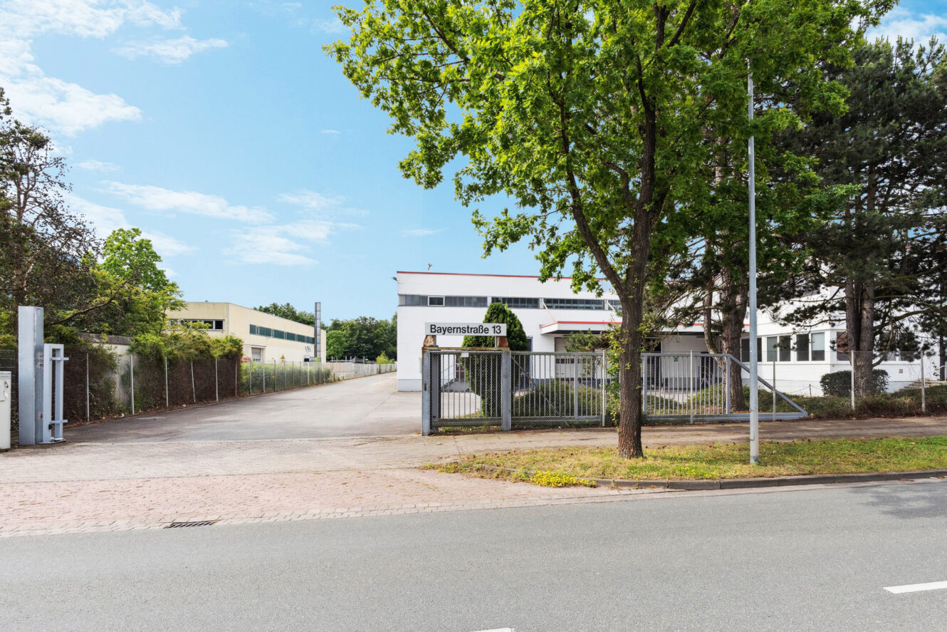Einfahrt zu einer Industrieanlage mit einem Schild mit der Aufschrift Bayernstraße 13, umgeben von Bäumen und Gebäuden, bei teilweise bewölktem Himmel.