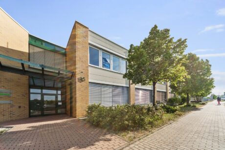 Ein modernes zweistöckiges Bürogebäude mit großen Fenstern, hellgelben Ziegelwänden und einem von Bäumen gesäumten Bürgersteig unter einem klaren blauen Himmel.
