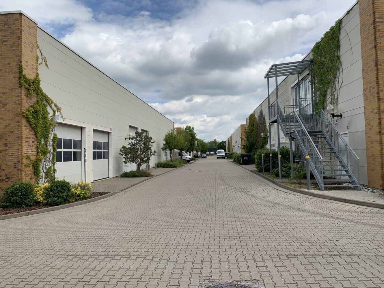 Industriegebiet mit zwei parallelen Lagergebäuden, einer Einfahrt in der Mitte, einer Metalltreppe auf der rechten Seite und etwas Grün an den Seiten unter einem bewölkten Himmel.