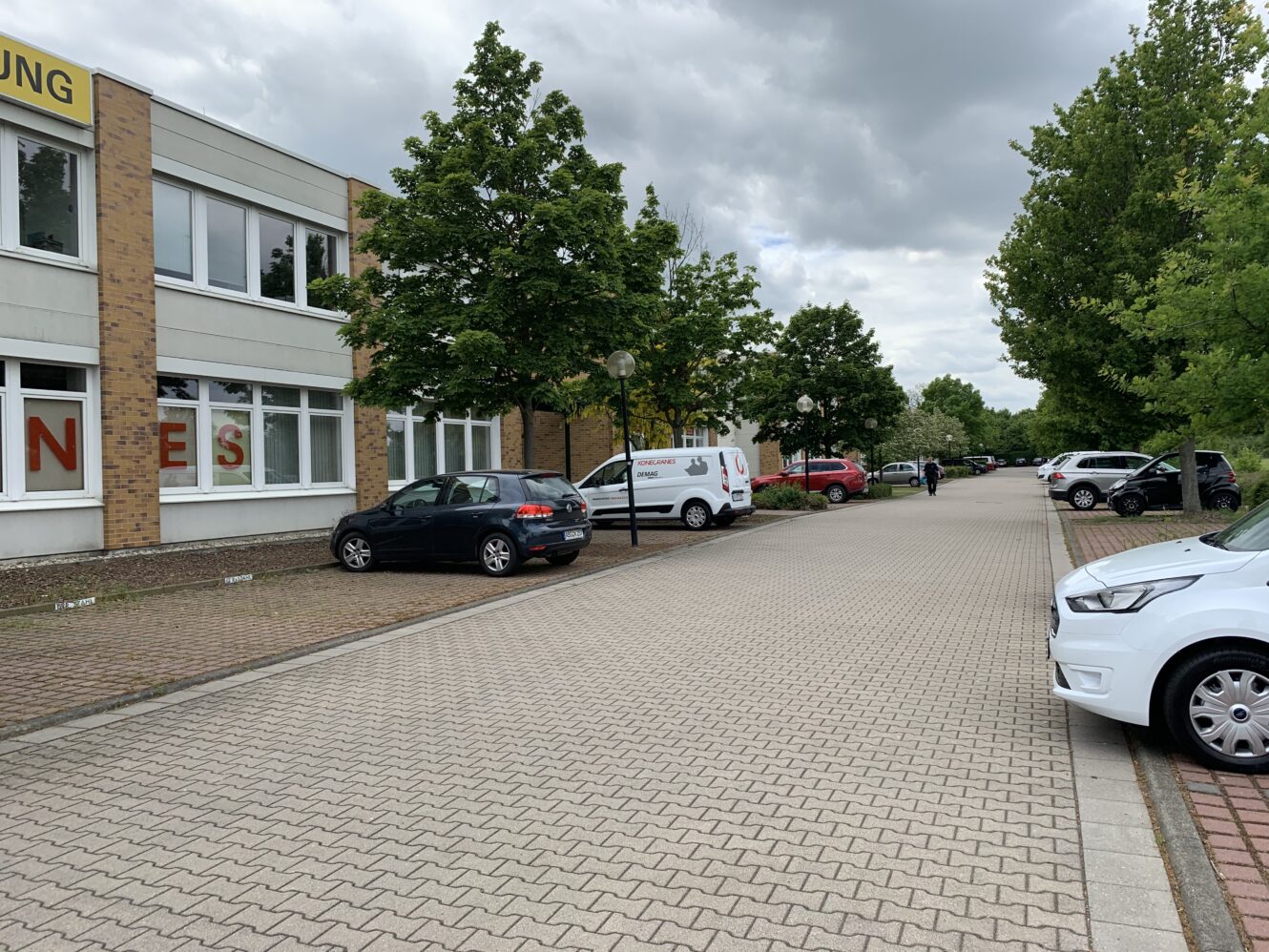 Ein Parkplatz mit mehreren Autos, die entlang einer gepflasterten Straße neben einem Gebäude geparkt sind, mit Bäumen auf beiden Seiten unter einem bewölkten Himmel.