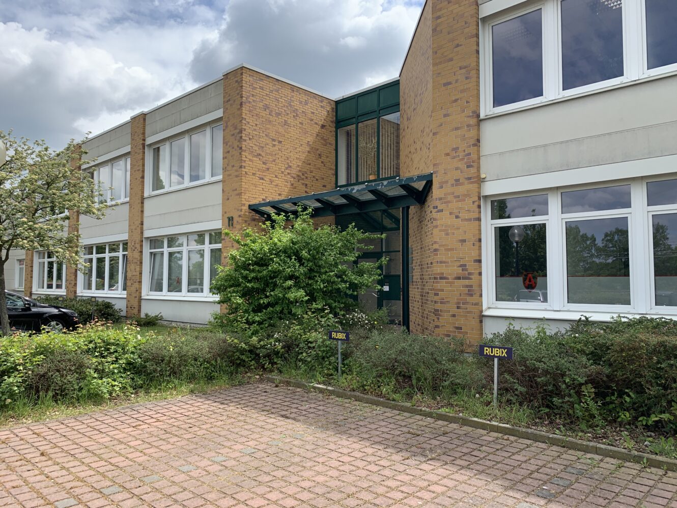 Zweistöckiges Bürogebäude mit weißem Backstein, großen Fenstern, Glaseingang, Grünanlagen und einem gepflasterten Parkplatz davor. Schilder mit der Aufschrift RUBYX sind sichtbar.