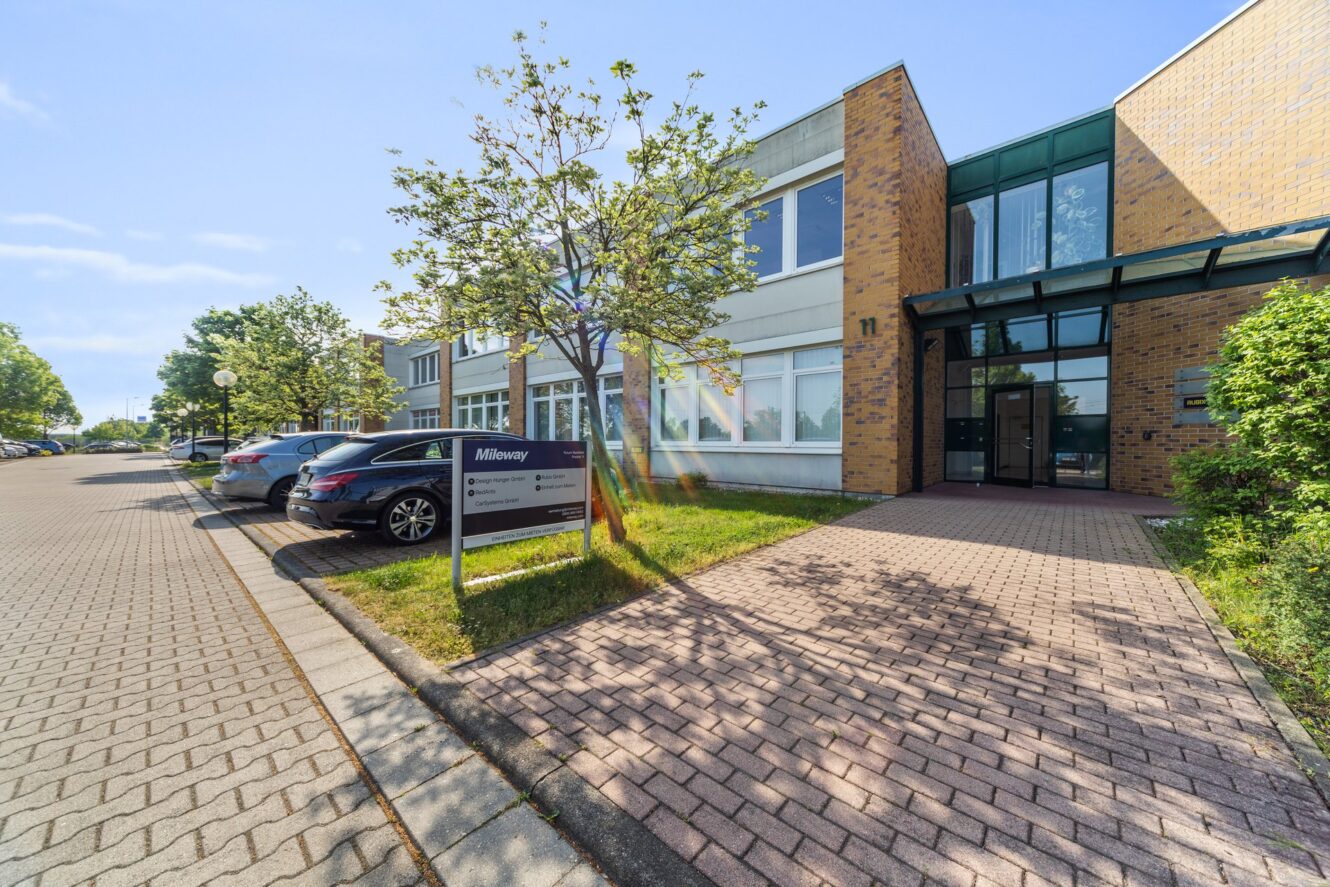 Außenansicht eines Bürogebäudes an einem sonnigen Tag mit mehreren geparkten Autos, einem Baum und einem Schild mit der Aufschrift "Miltenyi" neben dem Eingang.