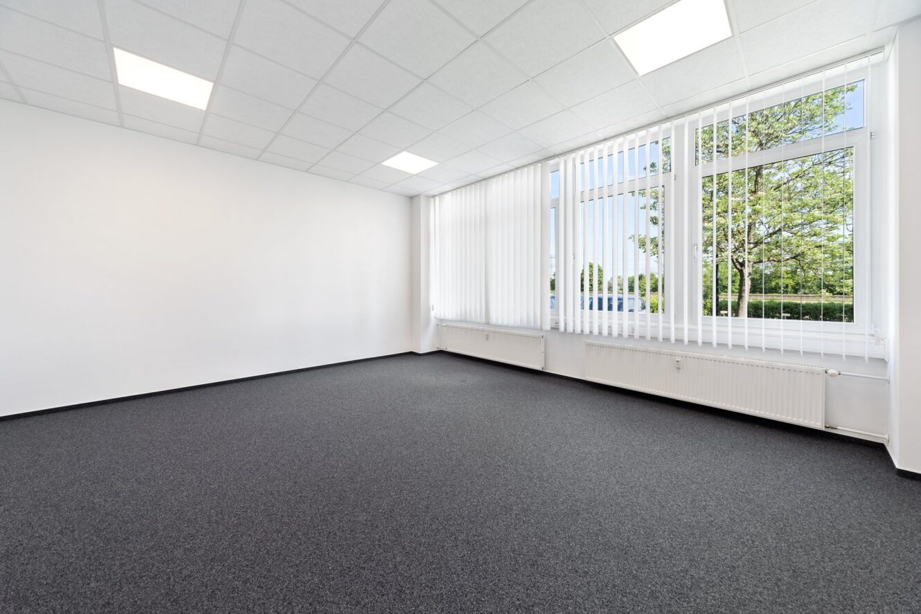 Leerer Büroraum mit weißen Wänden, großen Fenstern mit vertikalen Jalousien, Deckenleuchten und dunkelgrauem Teppichboden. Vor den Fenstern sind Bäume zu sehen.