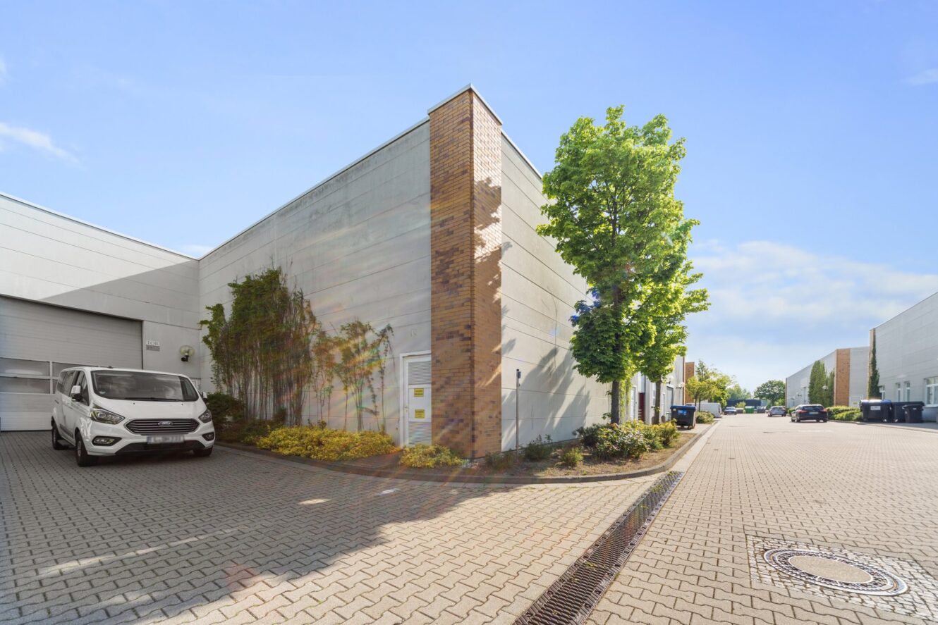 Ein weißes Lagerhaus mit einem vor dem Haus geparkten Lieferwagen, Ziegelsteinen und Bäumen entlang einer gepflasterten Straße unter einem strahlend blauen Himmel.