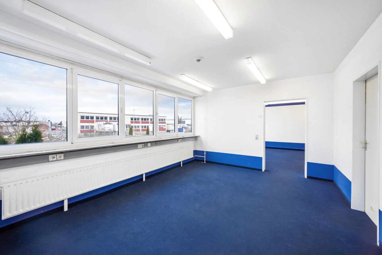 Leerer Büroraum mit blauem Teppich, weißen Wänden, fluoreszierenden Deckenleuchten, großen Fenstern und einem Heizkörper unter den Fenstern.