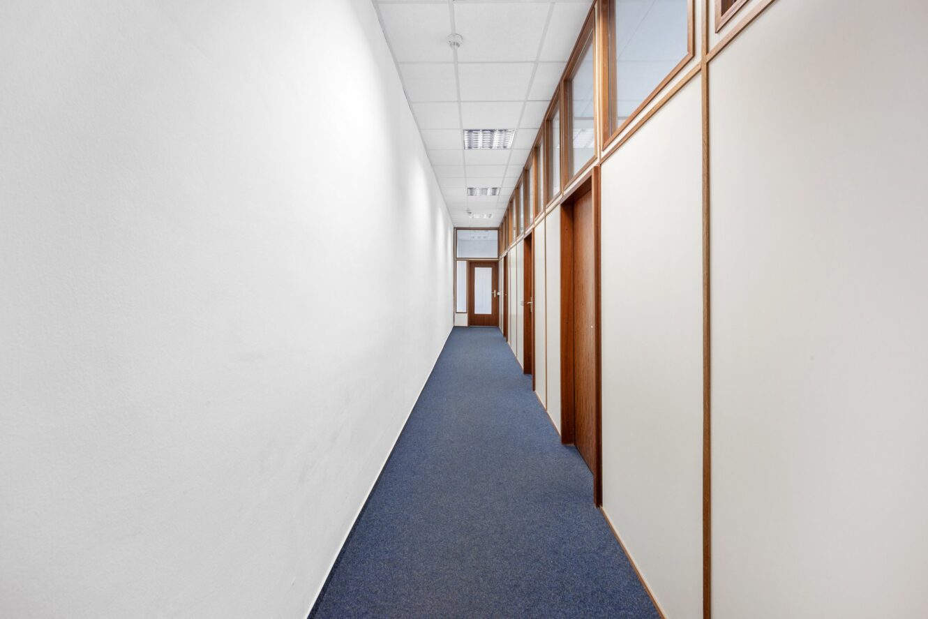 Büroflur mit weißen Wänden, blauem Teppich und holzgerahmten Glastrennwänden, die zu geschlossenen Türen am Ende führen. Deckenleuchten und Fliesen sind sichtbar.