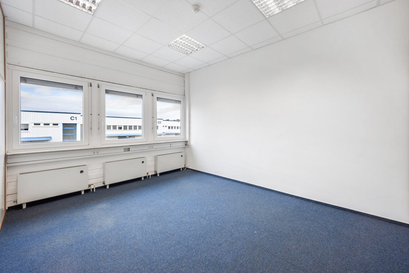 Ein leerer Büroraum mit weißen Wänden, blauem Teppich, drei großen Fenstern und Leuchtstoffröhrenbeleuchtung. Vor den Fenstern sind Industriegebäude zu sehen.
