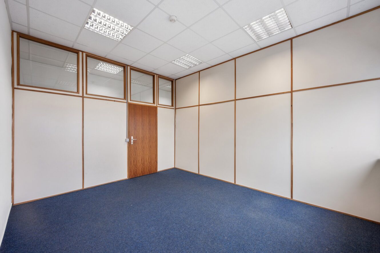 Ein leerer Büroraum mit blauem Teppich, weißen Wänden, Holzverkleidung, einer geschlossenen Holztür und Deckenleuchten.