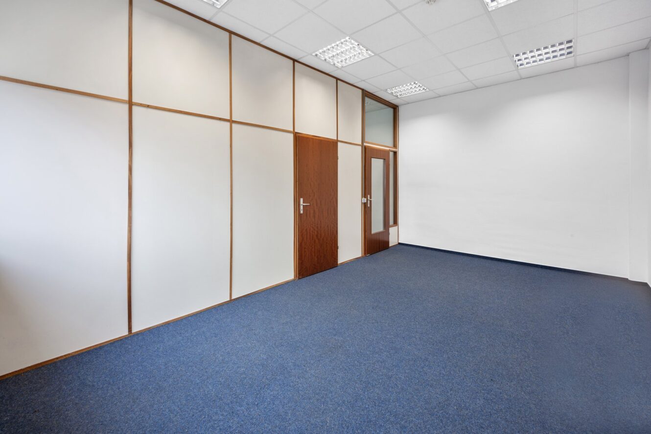 Leerer Büroraum mit blauem Teppich, weißen Wänden, holzgerahmten Glasscheiben, einer Holztür und einer fluoreszierenden Decke.