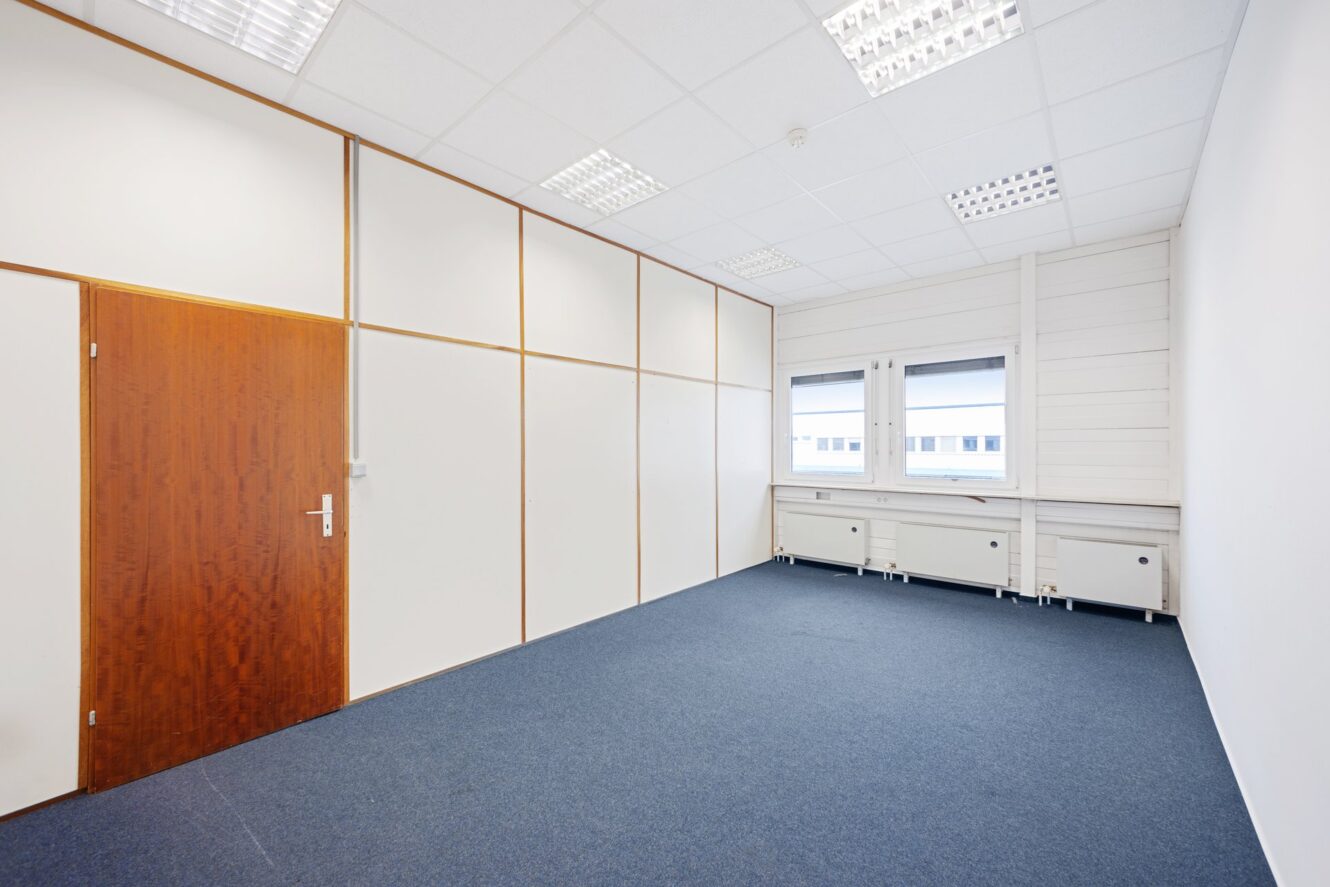 Leerer Büroraum mit blauem Teppich, weißen Wänden mit Holzverkleidung, einer geschlossenen Holztür und zwei Fenstern mit weißen Rahmen und Schränken darunter.