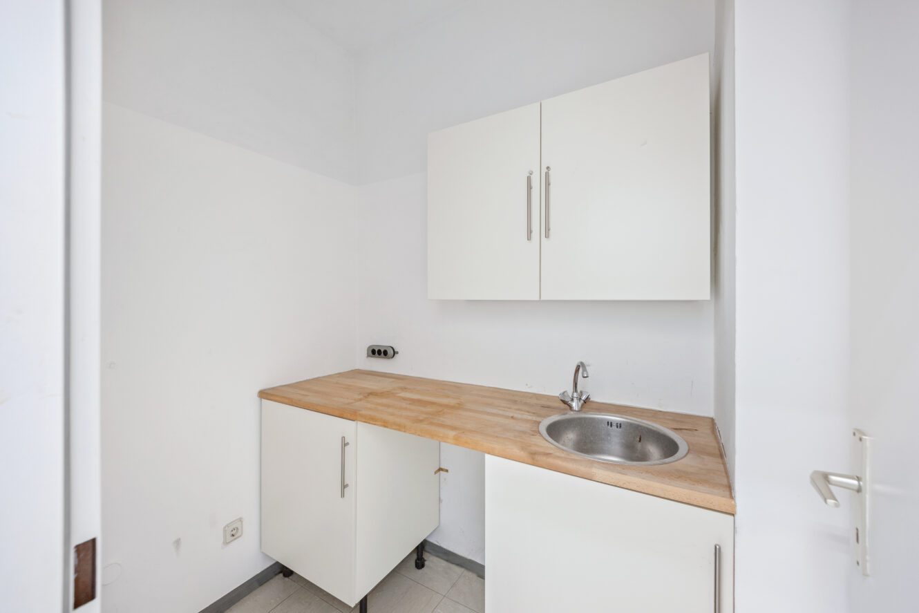 Kleine Küchenecke mit weißen Schränken, einer hölzernen Arbeitsplatte, einer runden Spüle und einem an der Wand befestigten Oberschrank; minimales Dekor und neutrale Farben.