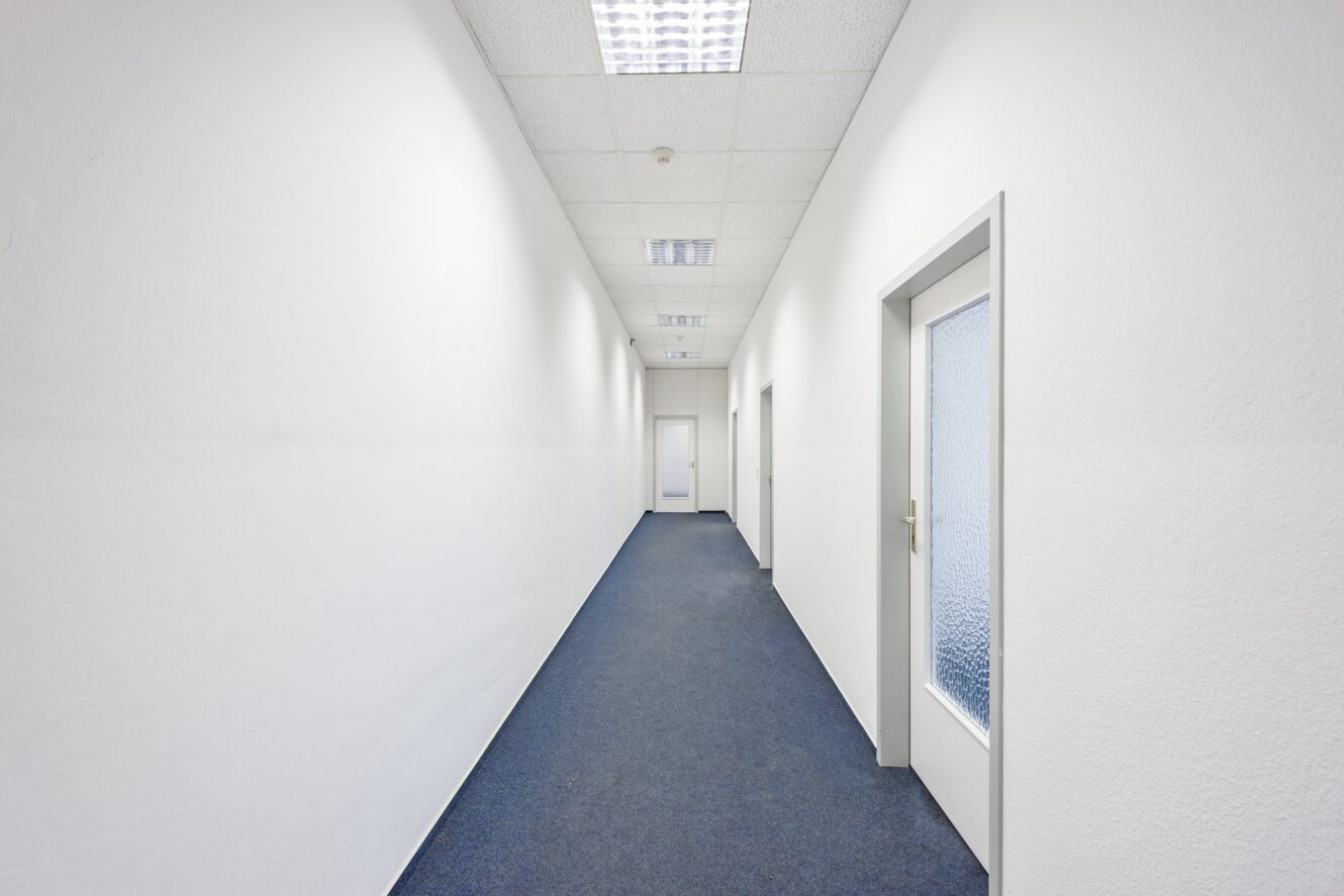 Ein langer, schmaler Flur mit weißen Wänden, blauem Teppich, Neonröhren und mehreren geschlossenen weißen Türen.