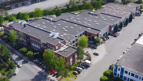 Luftaufnahme eines großen Industriegebäudes mit Flachdach, umgeben von Bäumen, parkenden Autos und angrenzenden Straßen.