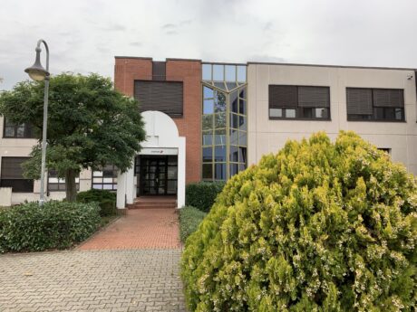 Ein modernes zweistöckiges Bürogebäude mit roten Ziegeln und beigen Wänden, großen Fenstern, einem grünen Busch davor und einem gepflasterten Weg, der zum Eingang führt.