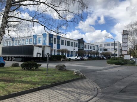Ein großes Industrie- oder Bürogebäude mit blauem und weißem Äußeren, ein geparkter Sattelschlepper, mehrere Autos, Bäume und ein Firmenschild unter einem teilweise bewölkten Himmel.