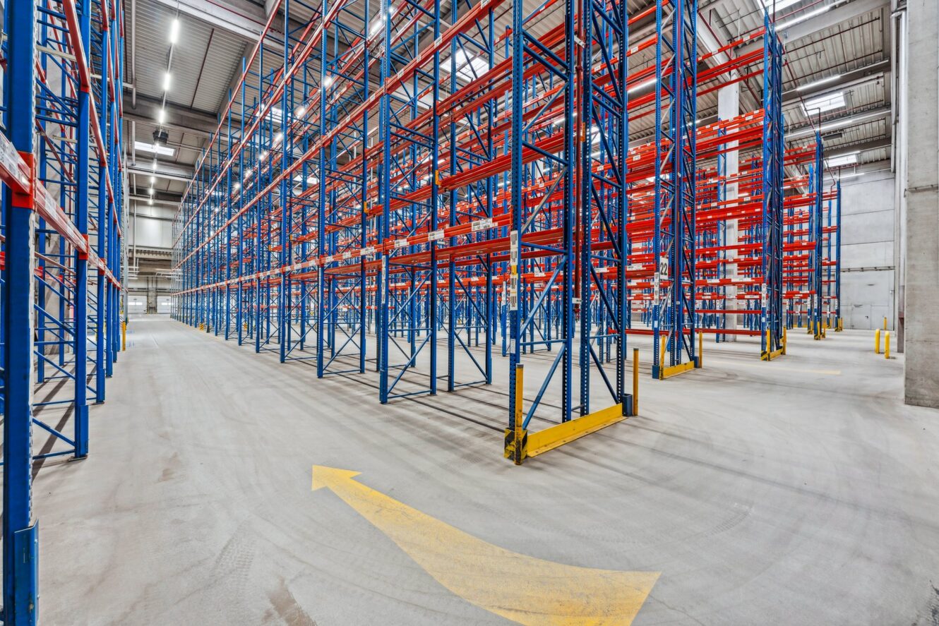 Leeres Lagerhaus mit hohen blauen und roten Metallregalen, Betonboden und einem auf den Boden gemalten gelben Richtungspfeil.