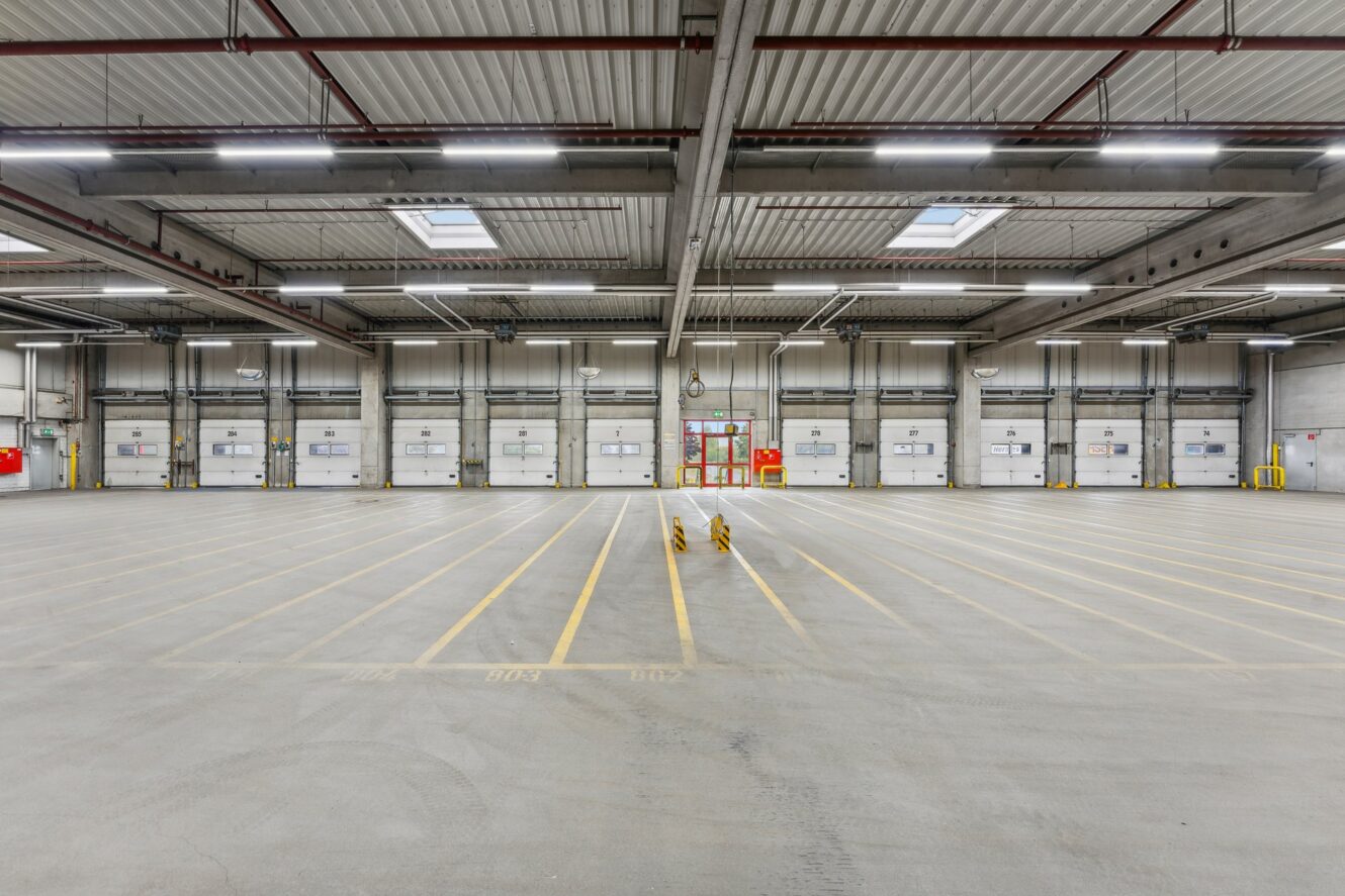 Leeres Lagerhaus mit hoher Decke, mehreren Laderaumtoren, gelb gestreiften Parklinien und Überkopfbeleuchtung.