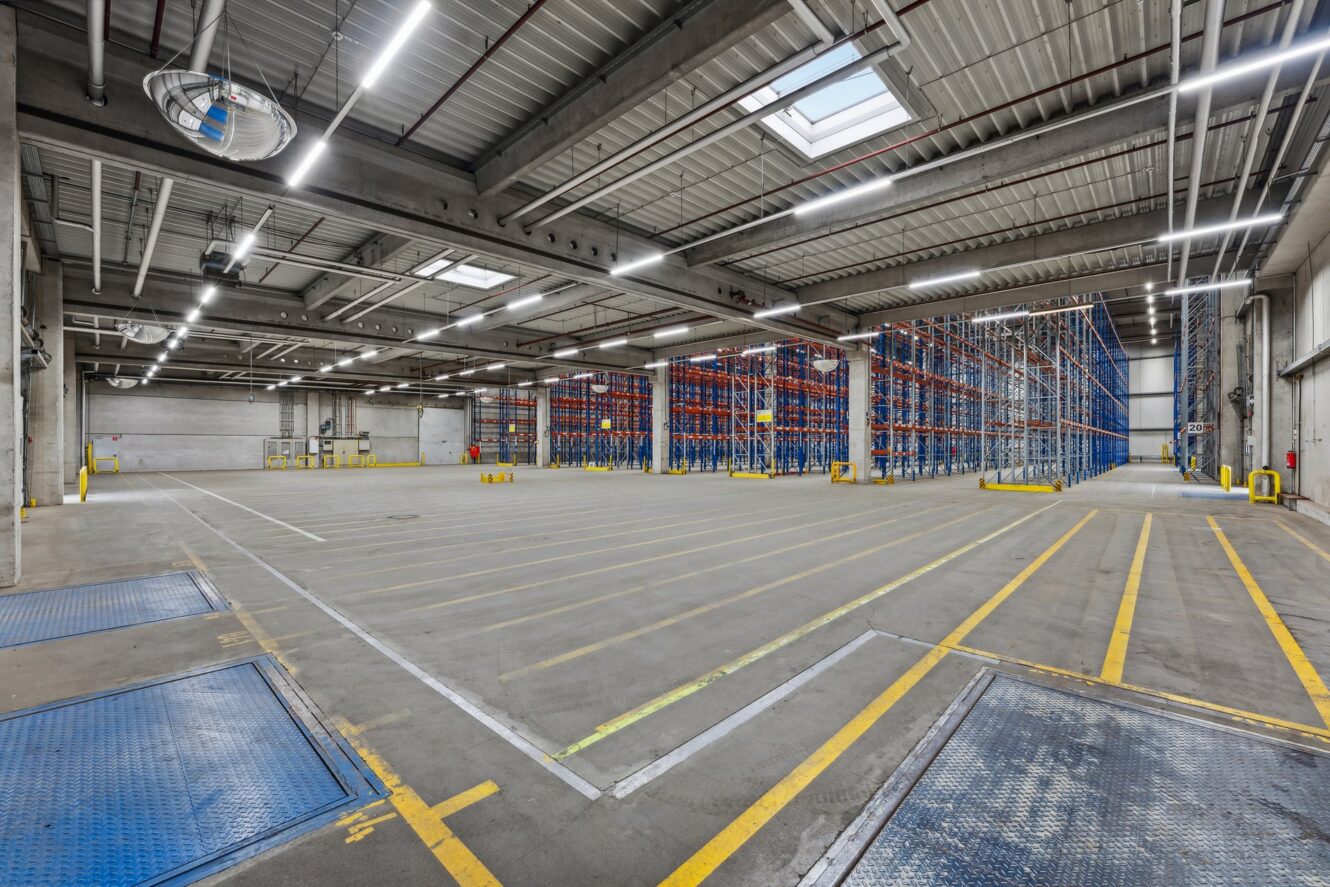 Leeres Lagerhaus mit hohen Decken, Metallregalen, Betonböden und markierten gelben Linien für Organisation und Fahrzeugwege. Helle Deckenbeleuchtung und Oberlichter sind vorhanden.